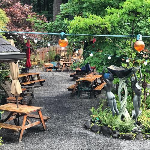 A garden area with wooden tables, string lights, and decorative sculptures amidst greenery and trees.