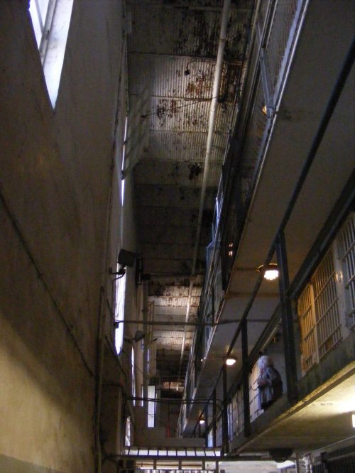 Interior view of a prison corridor with metal bars, high ceilings, and peeling paint, illuminated by overhead lights.
