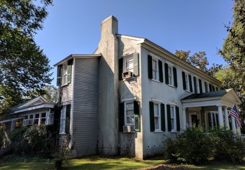 A two-story house with green shutters, a chimney, and an American flag, surrounded by trees and a clear blue sky.