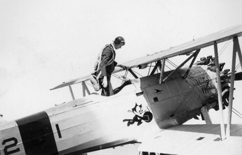 A pilot in a vintage biplane adjusts equipment on the wing, with a cartoon character painted on the fuselage.
