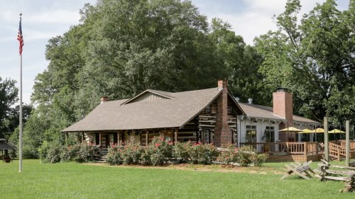 A charming log cabin-style house surrounded by greenery and flowers, with a flagpole in the foreground.