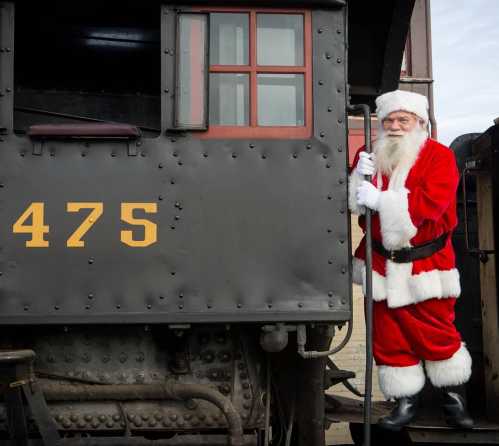 Santa Claus in a red suit stands on the steps of a vintage train, smiling and holding onto the railing.