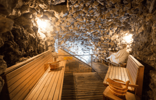 A serene stone sauna with wooden benches and a person sitting quietly in a robe, illuminated by soft lighting.