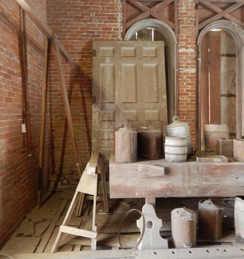 An old, rustic workshop with brick walls, wooden beams, a large door, and various containers on a wooden table.