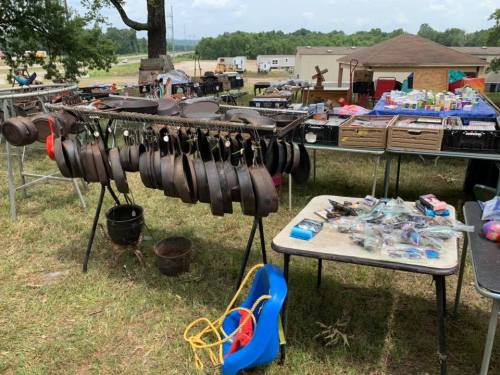 A flea market scene featuring hanging cast iron pans, tables with various items, and a grassy area.