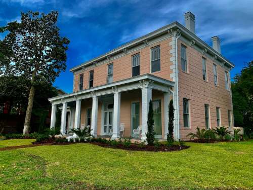 A large, pink two-story house with white columns, surrounded by green grass and palm trees under a blue sky.