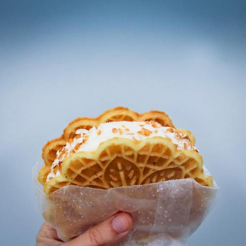 A hand holding a waffle sandwich filled with cream and topped with almond slices against a simple background.
