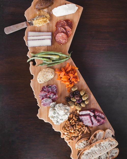 A charcuterie board shaped like New Jersey, featuring meats, cheeses, nuts, and vegetables on a wooden platter.