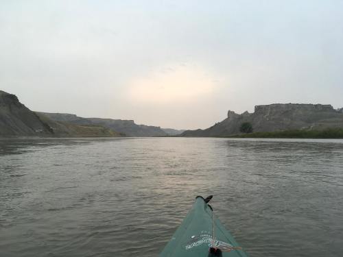 A kayak on a calm river, surrounded by rocky cliffs and a hazy sky at dusk.