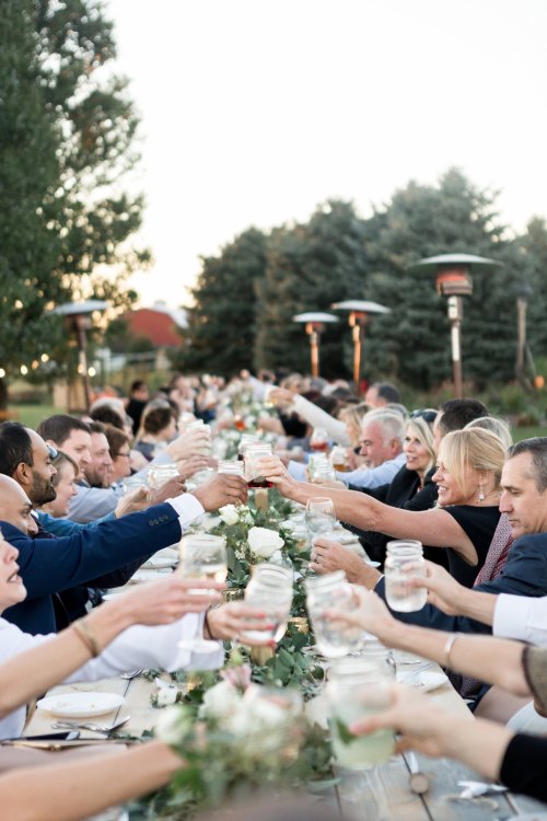A long outdoor table filled with people raising glasses in a toast, surrounded by greenery and warm lighting.