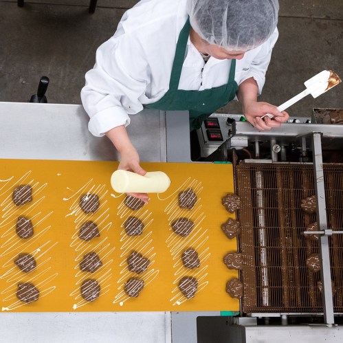 A worker drizzles white chocolate over freshly made chocolate confections on a production line.