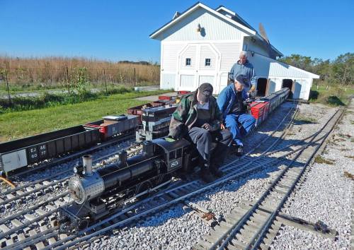 Two men sit on a miniature train track beside a small steam locomotive and a train station, surrounded by model train cars.