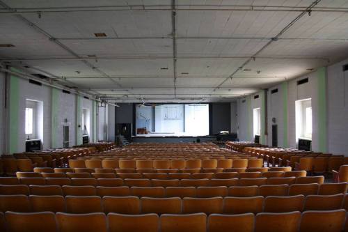 An empty auditorium with rows of wooden chairs facing a stage and large windows.