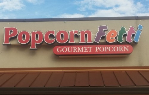 Sign for "PopcornFetti" featuring colorful letters, advertising gourmet popcorn against a blue sky backdrop.