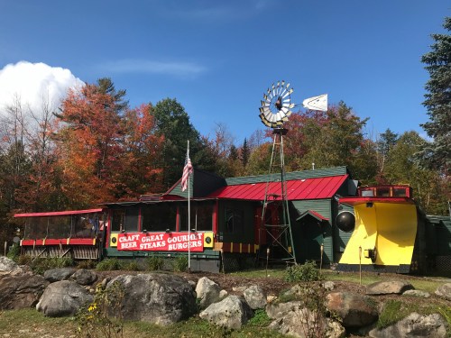 A colorful restaurant with a windmill, surrounded by autumn trees, featuring signs for burgers and craft beer.