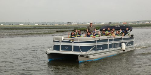 A group of people in life jackets smiles and waves from a pontoon boat on a calm waterway.