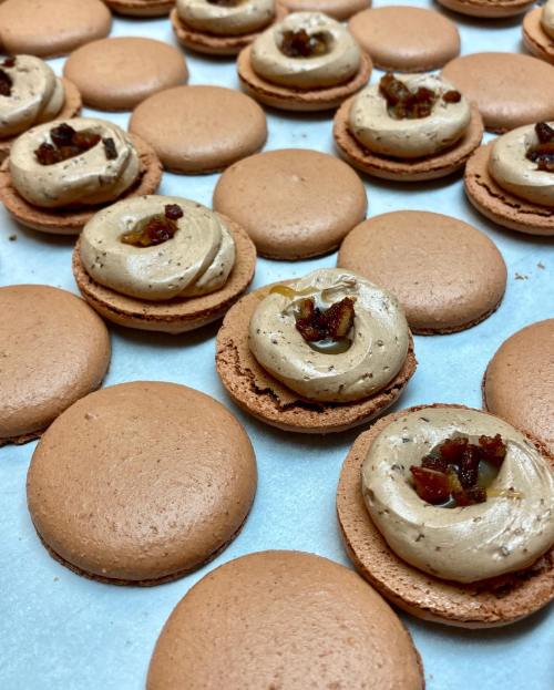 A close-up of chocolate macarons filled with creamy frosting and topped with bits of fruit, arranged on a baking sheet.