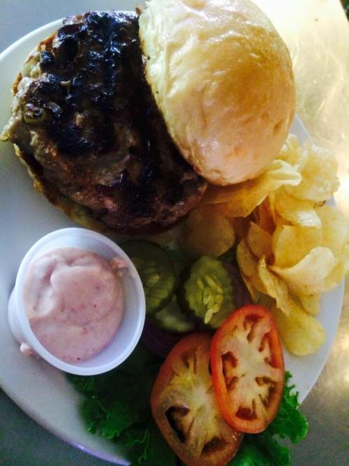 A grilled burger on a bun, served with lettuce, tomato, pickles, chips, and a side of dipping sauce.