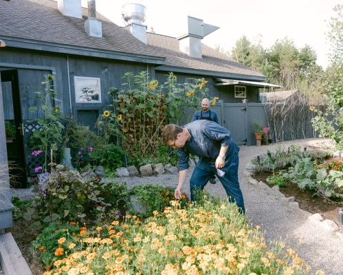 Two people tending to a vibrant garden filled with flowers and plants, with a house in the background.