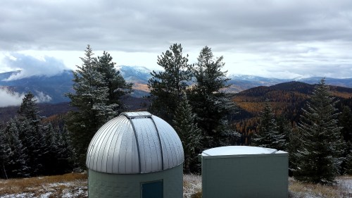 A scenic view of mountains and forests, featuring two observatory domes under a cloudy sky.