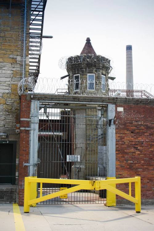 A prison entrance with a yellow gate, barbed wire, and a guard tower in the background.