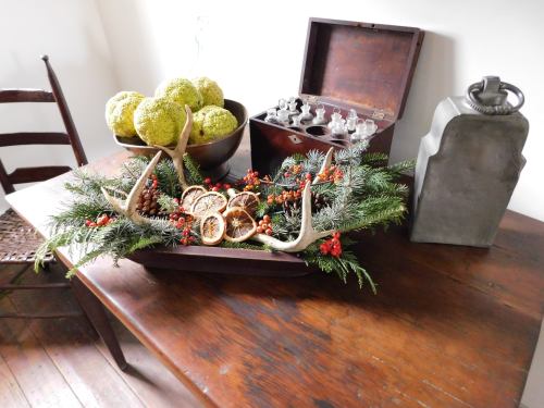 A rustic table displays a decorative arrangement with antlers, greenery, dried oranges, and a vintage box.