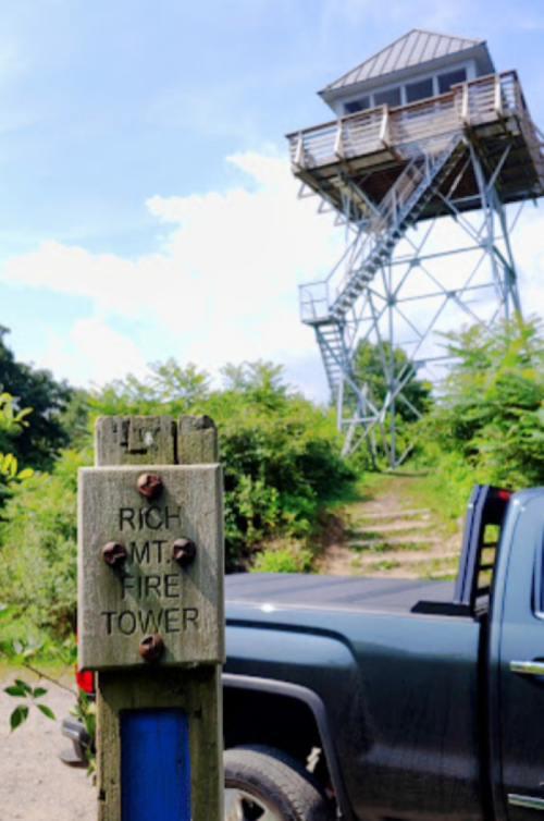 A wooden sign for "Rich Mt. Fire Tower" next to a parked truck, with a tall fire tower in the background and greenery around.