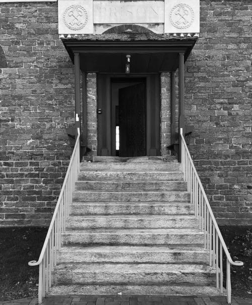 A stone building entrance with a set of stairs leading to a wooden door, framed by a red awning. Black and white image.