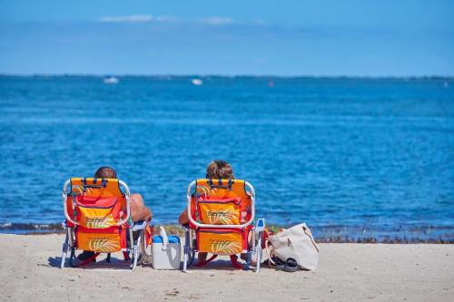 Two people relax in beach chairs by the water, enjoying a sunny day at the beach. A bag sits nearby on the sand.