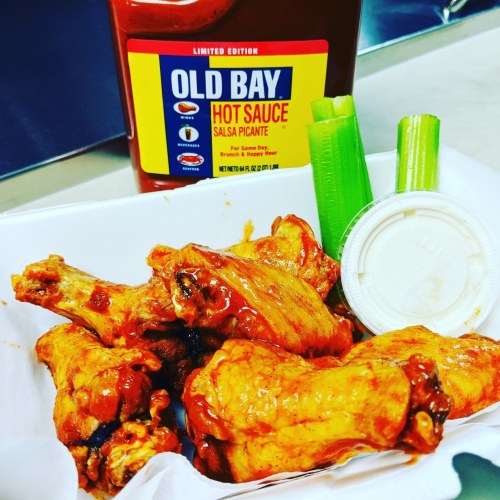 A plate of chicken wings coated in sauce, served with celery sticks and a cup of dipping sauce, alongside Old Bay hot sauce.