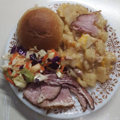 A plate of food featuring sliced ham, cheesy potatoes, a roll, and a colorful salad with various vegetables.