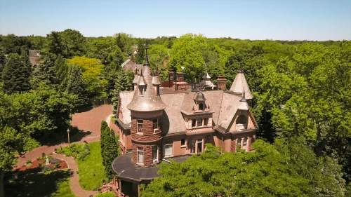 Aerial view of a large, ornate mansion surrounded by lush green trees and gardens on a sunny day.