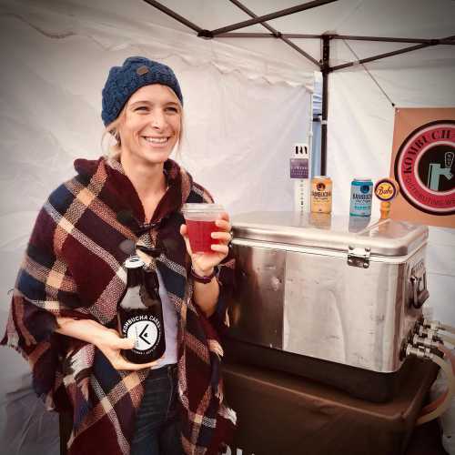 A smiling woman in a poncho holds a kombucha bottle and cup at a beverage stand with various drinks displayed.