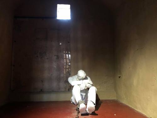 A solitary figure in a dimly lit cell, sitting against the wall with hands clasped, evoking a sense of confinement.