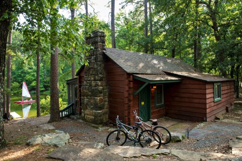 A rustic cabin surrounded by trees, with a stone chimney and bicycles parked outside, near a calm lake.