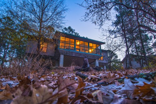 A modern house with large windows, surrounded by trees and fallen leaves, illuminated at dusk.