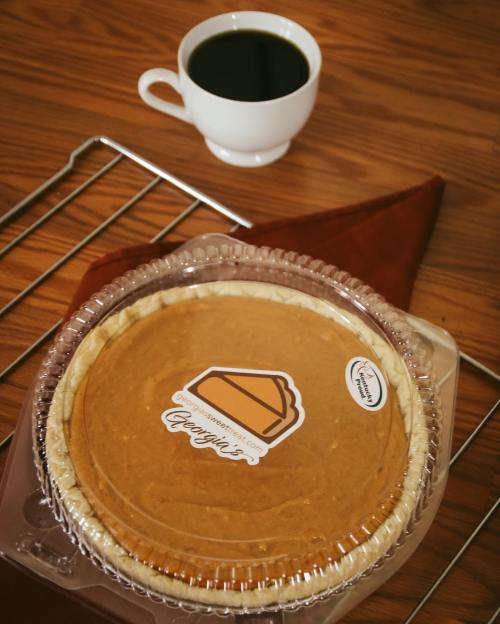 A pumpkin pie in a clear container with a label, next to a cup of coffee on a wooden surface.