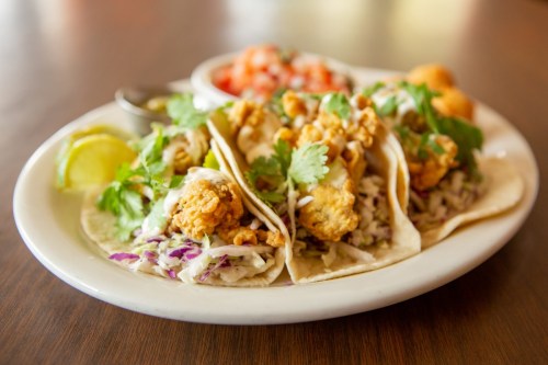 Three crispy fish tacos topped with cilantro and served with lime, salsa, and a side of coleslaw.
