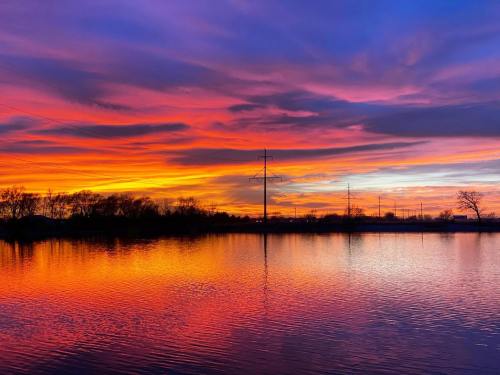 Vibrant sunset over a calm lake, with colorful clouds reflecting on the water and silhouettes of trees and power lines.