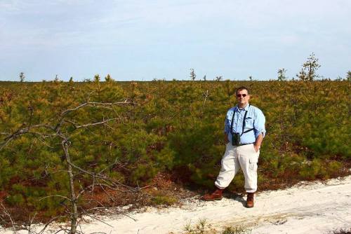 A person stands in a green field with pine trees, wearing a blue shirt and binoculars, against a clear sky.