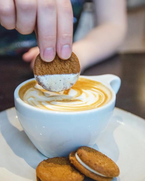 A hand dips a cookie into a frothy cup of coffee, with more cookies on the plate beside it.