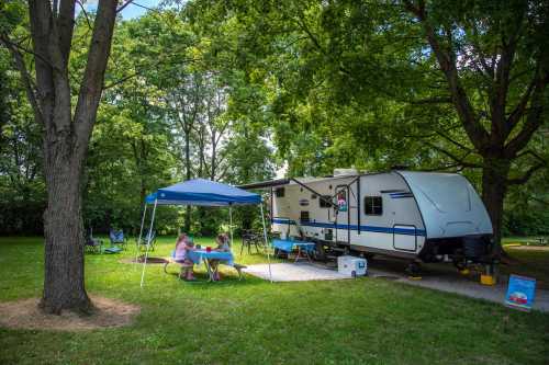 A camper trailer parked under trees, with two people sitting at a table enjoying the outdoors.