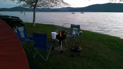 A lakeside campsite with two fishing chairs, a grill with a fire, and a bucket, surrounded by water and mountains.