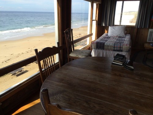 Cozy beachside room with a bed and wooden table, featuring large windows overlooking the sandy shore and ocean.