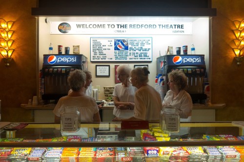 Four women in white shirts chat at the concession stand of the Redford Theatre, surrounded by snacks and drinks.