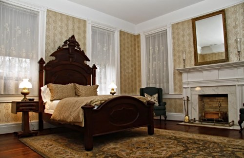 A cozy bedroom featuring a wooden bed, elegant decor, a fireplace, and vintage wallpaper. Soft lighting enhances the ambiance.
