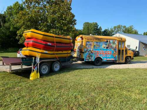 A colorful bus parked next to a trailer loaded with yellow and red rafts, set in a grassy area with trees in the background.