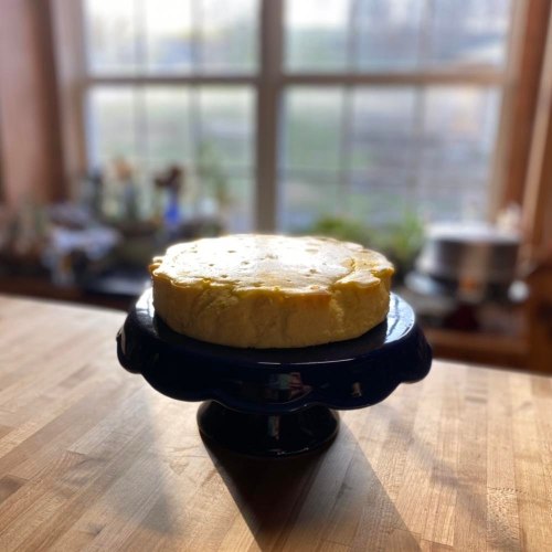 A cheesecake on a blue cake stand, set on a wooden table with a sunny window in the background.