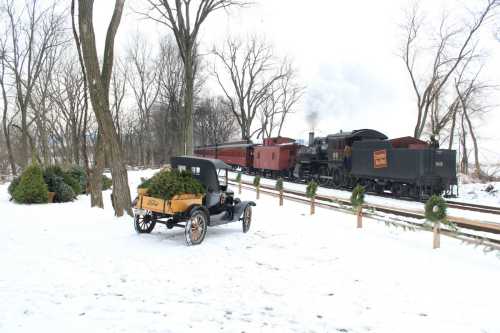 A vintage car parked in the snow with a steam train passing by in the background, surrounded by bare trees.