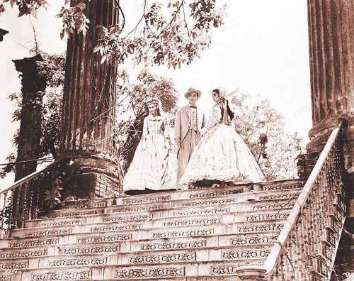 A sepia-toned image of three people in vintage clothing walking down ornate stairs surrounded by greenery.